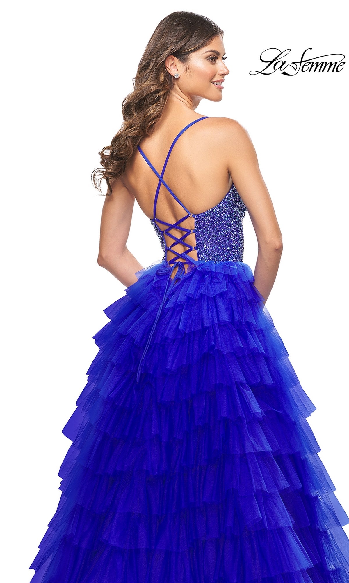 Woman in the La Femme Long Ruffled Prom Ball Gown 32002, featuring a royal blue beaded bodice and crisscross lace-up back, stands looking over her shoulder in this stunning prom dress.